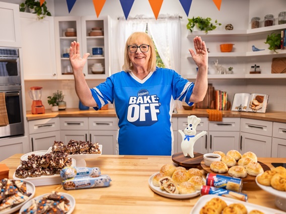 Donna Kelce and the Pillsbury Doughboy standing in front of a Game Day spread including Pillsbury rolls and cookie dough, celebrating the 53rd Pillsbury Bake-Off Contest.